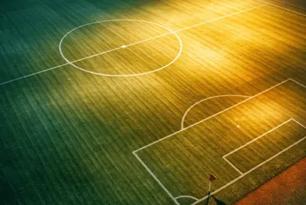 Empty floodlit football pitch at dusk with geometric shadows and national colour-toned lighting; overhead wide-angle view.