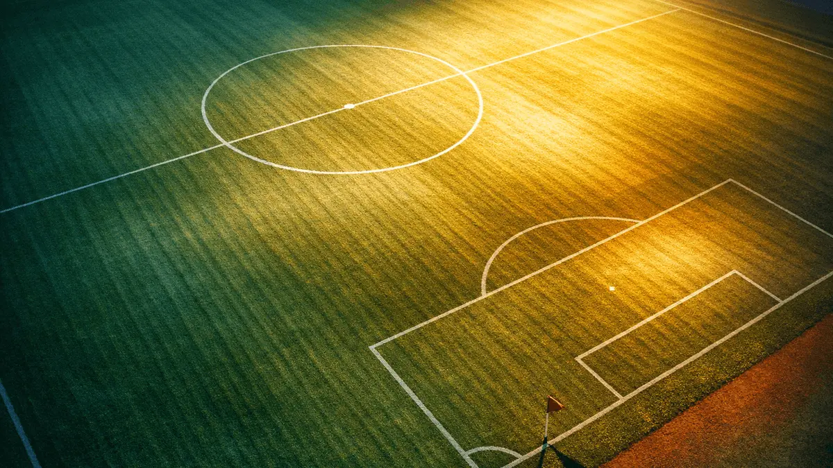 Empty floodlit football pitch at dusk with geometric shadows and national colour-toned lighting; overhead wide-angle view.