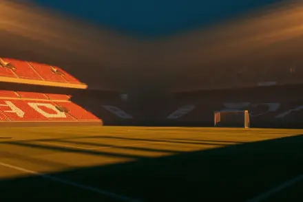 Floodlit football stadium at dusk with red and white seats and dramatic shadows on empty pitch.