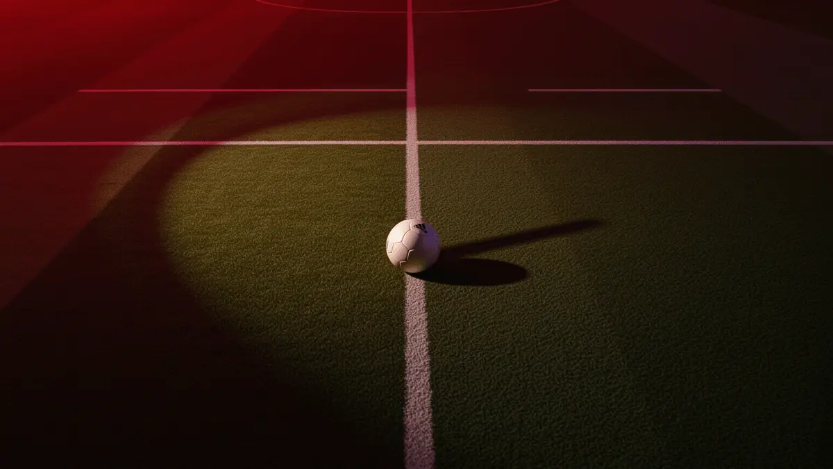 Empty floodlit football pitch at dusk with match ball on centre spot, lit in red and white tones.