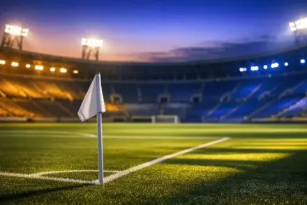 Empty floodlit stadium corner view with Cape Verde-coloured light accents and dramatic evening shadows on the pitch.