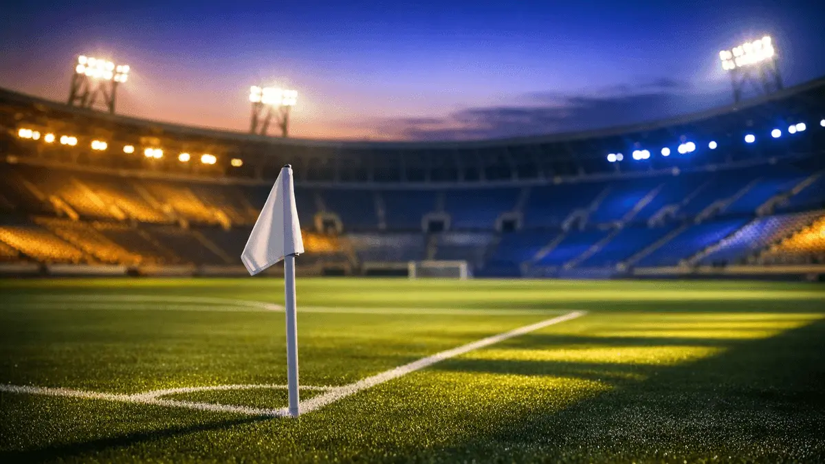 Empty floodlit stadium corner view with Cape Verde-coloured light accents and dramatic evening shadows on the pitch.