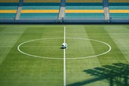 Elevated stadium view with turquoise and yellow pitch markings, match ball on centre spot, floodlit pylons casting shadows.