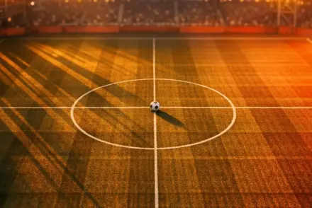 Aerial view of floodlit football pitch at golden hour with match ball on centre spot, red and gold lighting.