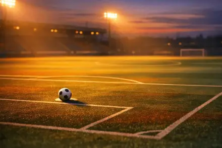 Floodlit stadium pitch at dusk with match ball at penalty spot, geometric markings in warm golden light, corner-flag angle view.