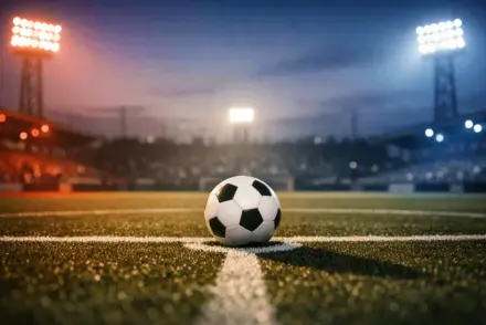 Floodlit football pitch at dusk with match ball on centre spot, red and blue stadium lighting, empty stands in background.