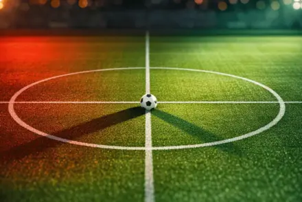 Aerial view of floodlit football pitch at dusk with white ball on centre spot, red and green lighting accents.
