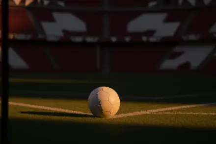 Floodlit football pitch with match ball at penalty spot under spotlight; maroon and white national colours in stand sections; dusk mood.