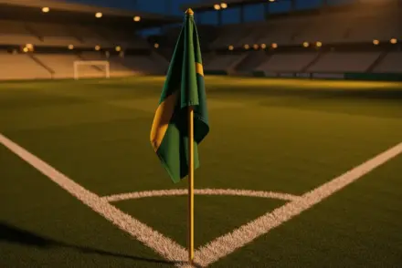 Aerial view of floodlit pitch at dusk with corner flag, emerald and gold light cast, dramatic shadows, no text.