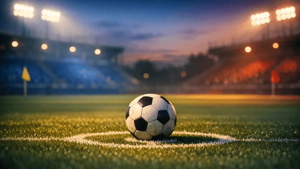 Floodlit football pitch with match ball on penalty spot, blue and red stadium lighting, evening match preview atmosphere.