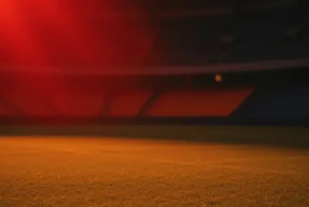 Floodlit stadium pitch at dusk with dramatic shadow patterns, red and orange lighting, empty stands.