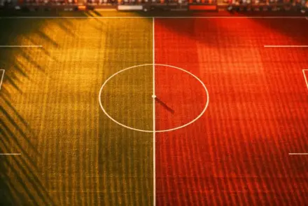 Overhead floodlit football pitch with centre circle and halfway line, red and white strategic lighting, golden-hour shadows, broadcast gantry angle.