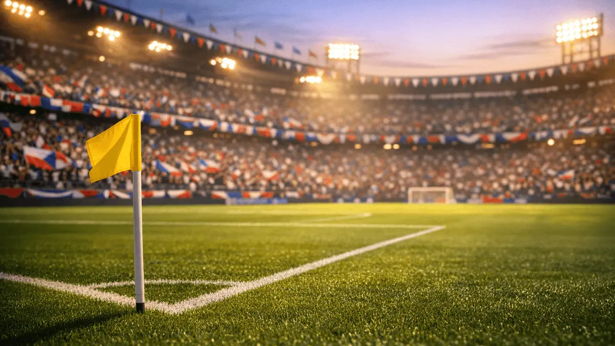 Stadium lower-bowl view with packed stands, flag bunting, and floodlit pitch during pre-match buildup.