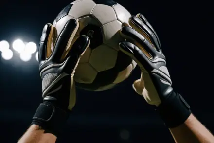 Goalkeeper's gloved hands making a decisive save in dramatic stadium lighting