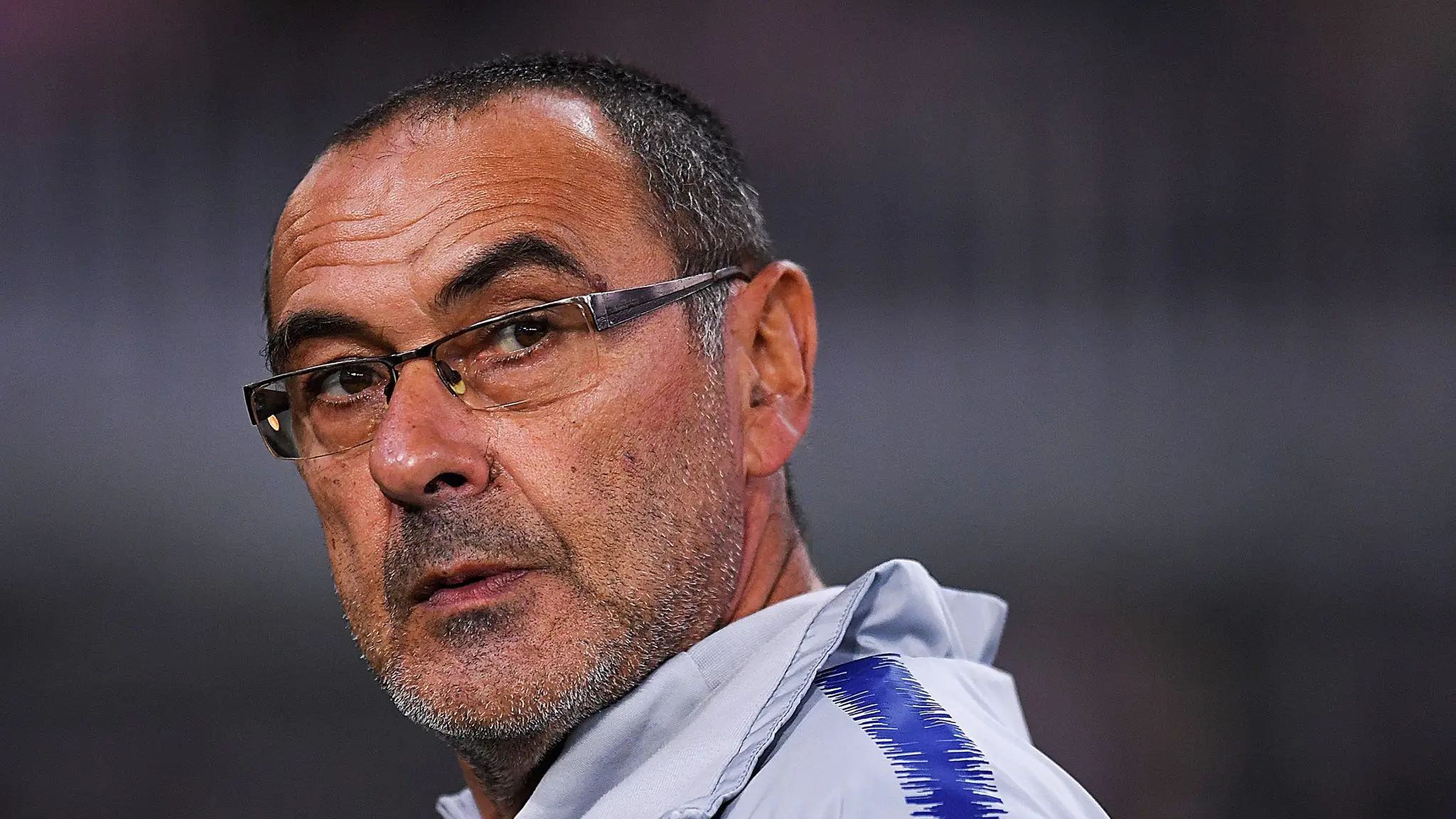 Close de Maurizio Sarri com expressão séria usando óculos e jaqueta cinza.