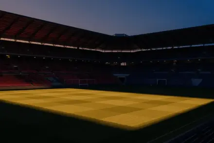 Floodlit football stadium at dusk with dramatic shadow patterns and golden-hour lighting, empty pitch.