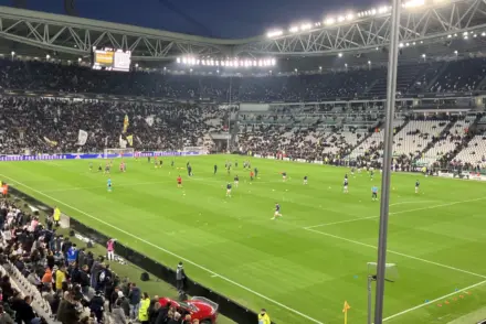A wide shot of the Allianz Stadium in Turin with players warming up on the pitch and fans in the stands.